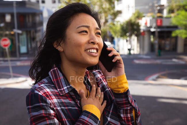 Side view close up of a mixed race woman with long dark hair out and about in the city streets during the day, talking on the smartphone, wearing a checked shirt and walking in a city street with buildings in the background. — Stock Photo