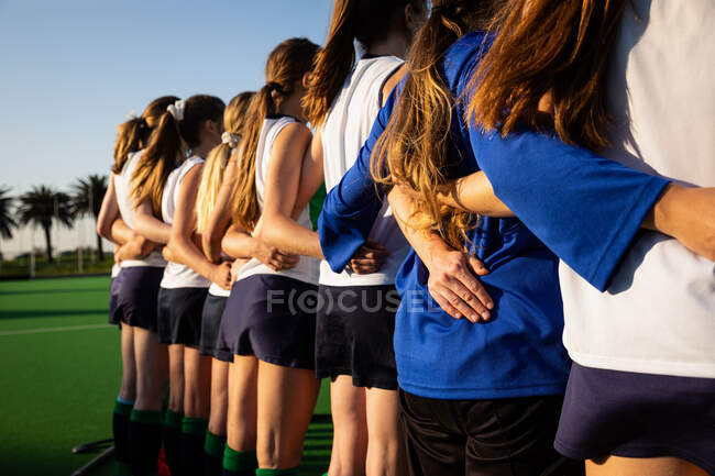 Visão traseira de um grupo de jogadoras de hóquei em campo caucasianas, preparando-se antes de um jogo, de pé em uma fileira abraçando, em um dia ensolarado — Fotografia de Stock