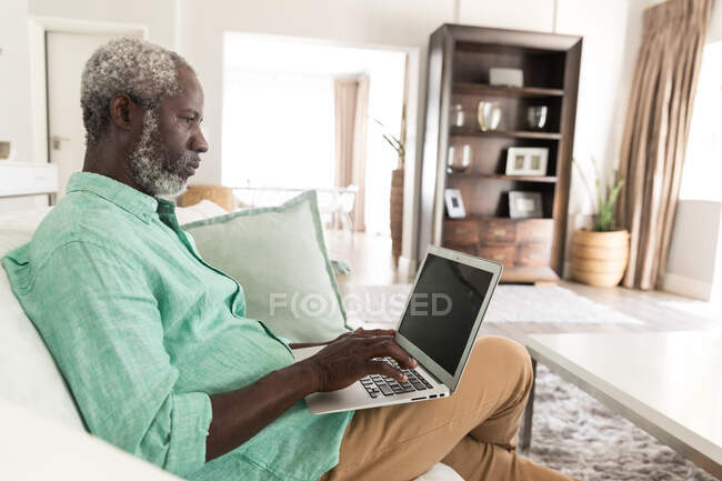 Старший афроамериканец проводит время дома, социальное дистанцирование и самоизоляция в карантинной изоляции во время эпидемии коронавируса, сидит на диване и использует ноутбук — стоковое фото
