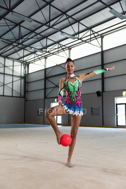 Vue de face d'une adolescente gymnaste mixte jouant au gymnase, faisant de l'exercice avec une balle rouge, debout avec la balle entre ses pieds, portant un justaucorps multicolore — Photo de stock