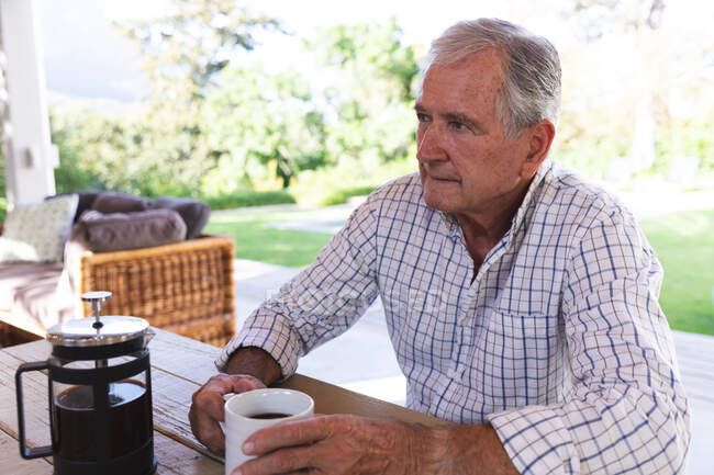 Счастливый пенсионер-европеец дома в саду возле своего дома в солнечный день, сидя на скамейке, держа чашку кофе, глядя в сторону и улыбаясь, самоизолируясь во время пандемии коронавируса — стоковое фото