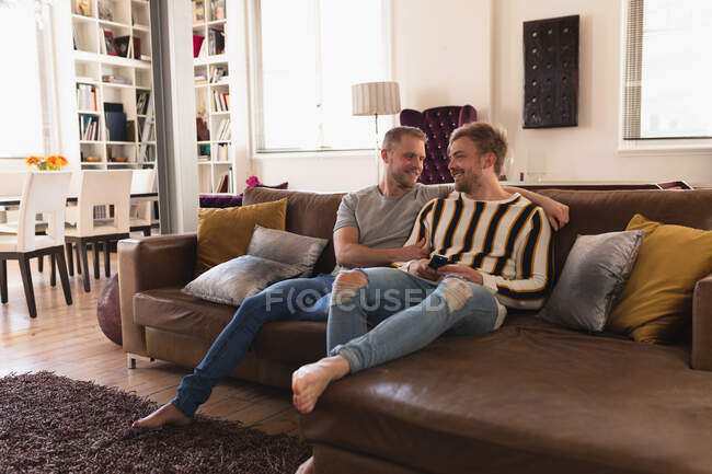Передній погляд Кавказької пари чоловіків відпочиває вдома, сидячи на дивані, обіймаючи, спілкуючись за допомогою смартфона. — стокове фото