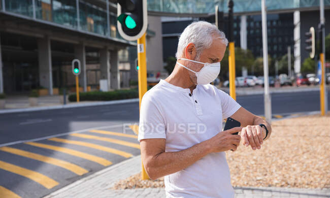 Homme caucasien âgé dans les rues de la ville pendant la journée, portant un masque facial contre le coronavirus, covid 19, à l'aide d'un smartphone et smartwatch. — Photo de stock