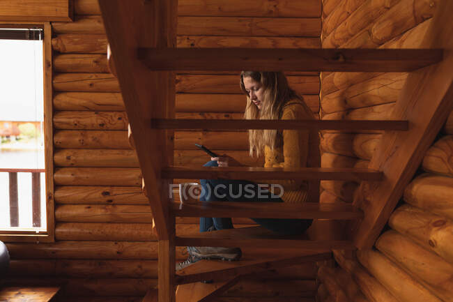 На вигляд кавказька жінка добре проводить час, подорожуючи горами, сидячи на сходах у хатині, користуючись смартфоном. — стокове фото
