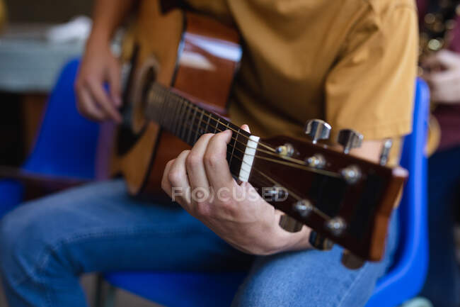 Front view mid section of musician teenage boy with yellow t-shirt sitting and playing an acoustic guitar in a high school — Stock Photo