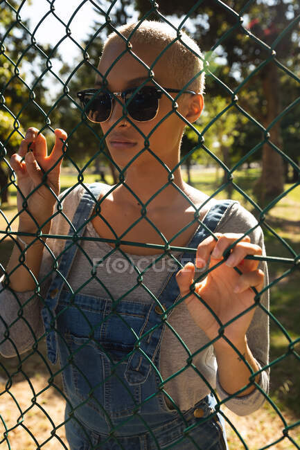 Смешанная расовая альтернативная женщина с короткими светлыми волосами в городе в солнечный день, в солнцезащитных очках и джинсовых комбинезонах, смотрящая через забор звеньев цепи. Городская независимая женщина на ходу — стоковое фото