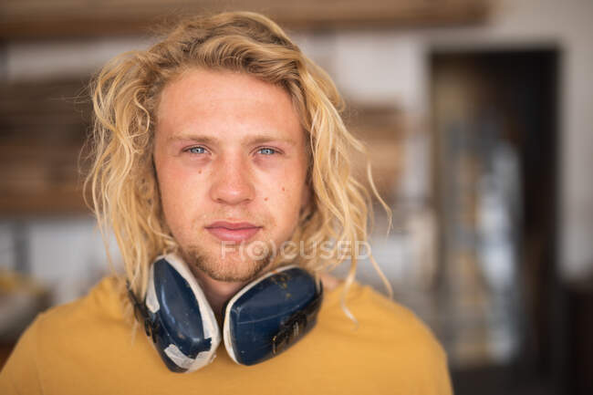 Porträt eines kaukasischen Surfbrettmachers mit langen blonden Haaren, der in seinem Studio steht, schützende Kopfhörer trägt und in die Kamera schaut. — Stockfoto
