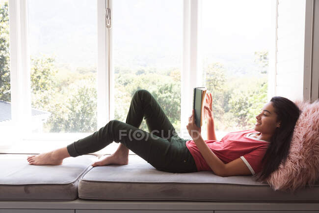Frau mit gemischter Rasse, die während der Coronavirus-Epidemie 19 in Quarantäne lebt und sich zu Hause selbstisoliert und sozial distanziert, auf Fensterbank liegend und im Wohnzimmer ein Buch liest. — Stockfoto