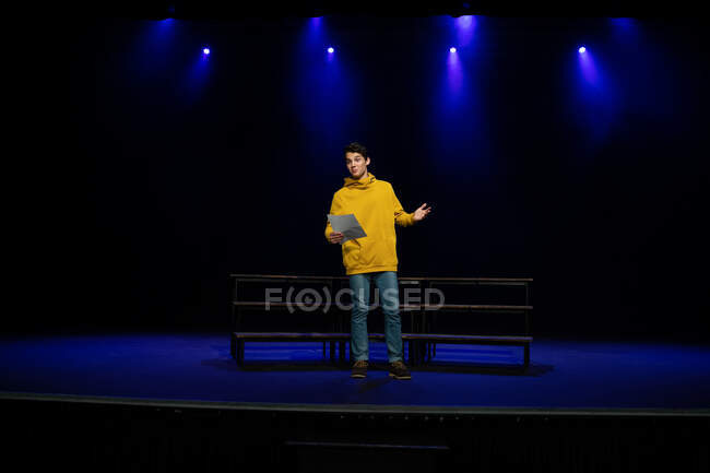 Вид спереди кавказского подростка, стоящего на сцене с сценарием в пустом школьном театре во время репетиций спектакля — стоковое фото
