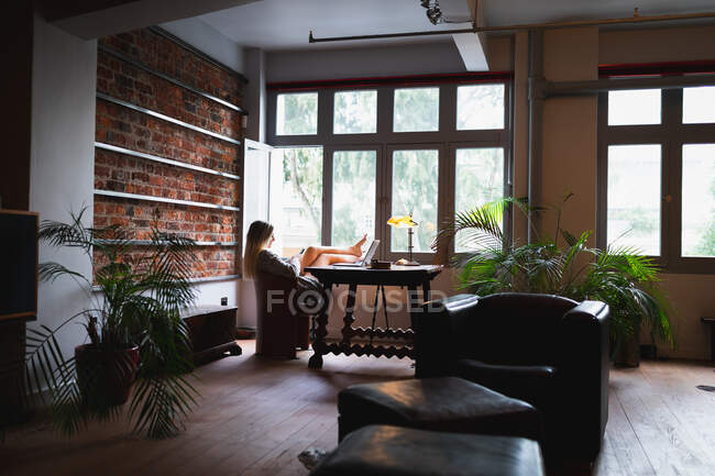 Autoaislamiento en cuarentena de encierro. vista lateral de una joven mujer caucásica, sentada en su oficina en casa, usando una computadora portátil mientras trabaja. - foto de stock