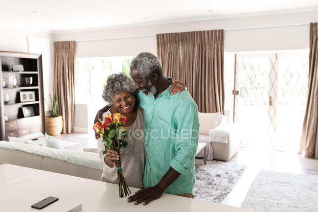 Старша афроамериканська пара проводить час удома разом, спілкуючись і самоізолюючись під час карантинної блокування під час епідемії коронавірусу (19), обіймаючи і посміхаючись, жінка тримає букет квітів. — стокове фото