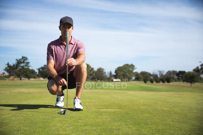 Vue de face d'un homme caucasien sur un terrain de golf par une journée ensoleillée, agenouillé, tenant un club de golf — Photo de stock