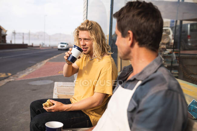 Dois fabricantes de pranchas de surf caucasianos fazendo uma pausa durante o trabalho em seu estúdio, sentados em um banco e interagindo enquanto bebem café e fazem um lanche . — Fotografia de Stock