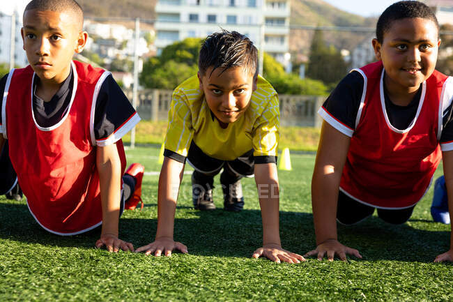 Vista frontale da vicino di un gruppo multietnico di giocatori di calcio maschili che fanno press up di fila su un campo da gioco al sole durante una sessione di allenamento di calcio — Foto stock