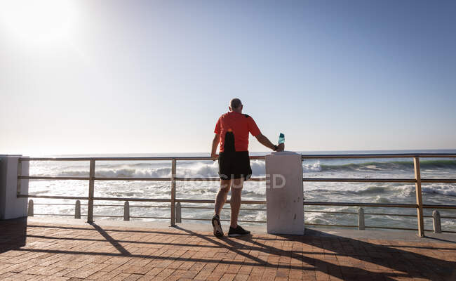 Vista trasera de un hombre mayor caucásico maduro haciendo ejercicio en un paseo marítimo en un día soleado con cielo azul, tomando un descanso, de pie, sosteniendo una botella de agua - foto de stock