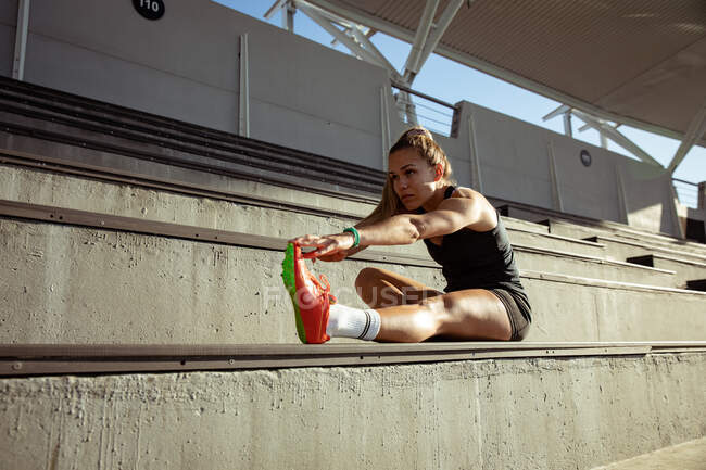 Side view of a Caucasian female athlete practicing at a sports stadium, sitting in the stands and stretching, holding her foot — Stock Photo