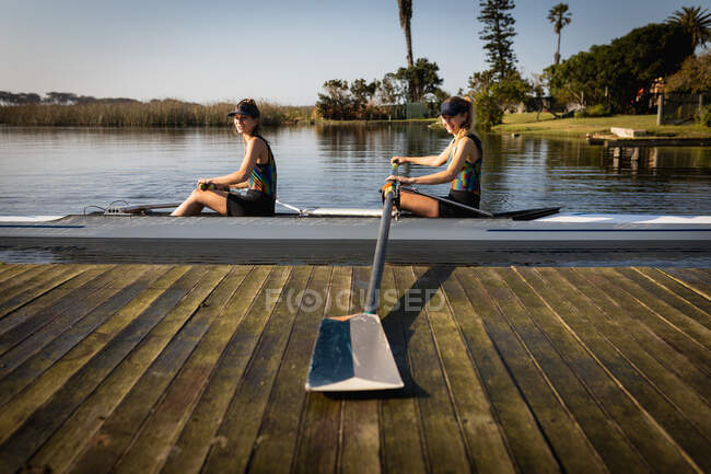Vista laterale di due vogatrici caucasiche da una squadra di canottaggio che si allenano sul fiume, sedute in un guscio da corsa sull'acqua e che si allontanano dal molo con un remo, sorridendo al sole — Foto stock
