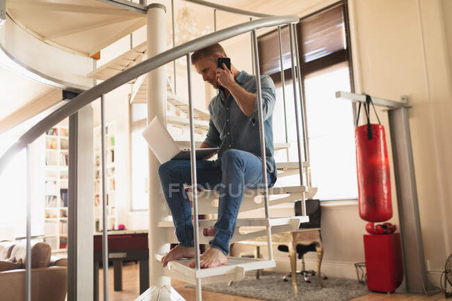 Front view of a young Caucasian man spending time at home, sitting on the stairs, talking on his smartphone and working on his laptop — Stock Photo