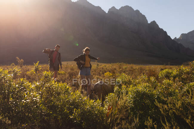 Vista laterale di una coppia caucasica che si diverte durante una gita in montagna, camminando su un campo sotto le montagne, in una giornata di sole — Foto stock