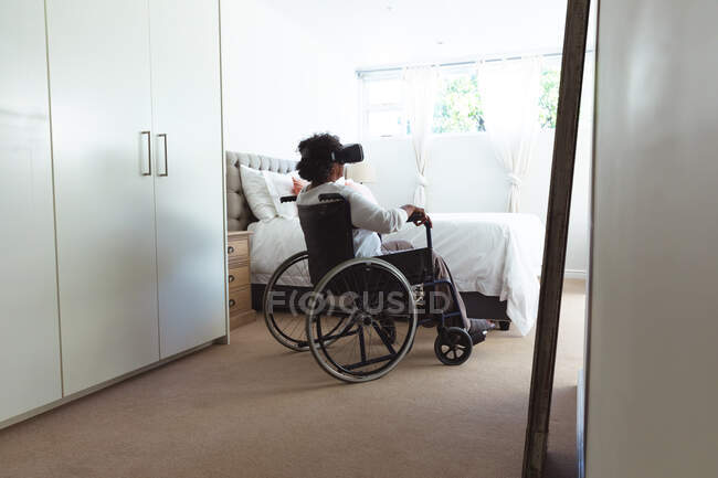 Mujer de raza mixta mayor disfrutando de su tiempo en casa, distanciamiento social y aislamiento en cuarentena, sentada en una silla de ruedas, con gafas vr - foto de stock