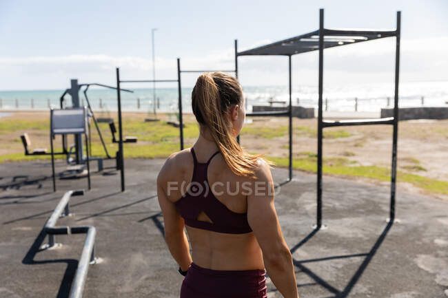 Vista posteriore di una donna caucasica sportiva con lunghi capelli scuri che si esercita in una palestra all'aperto durante il giorno, guardando il mare. — Foto stock