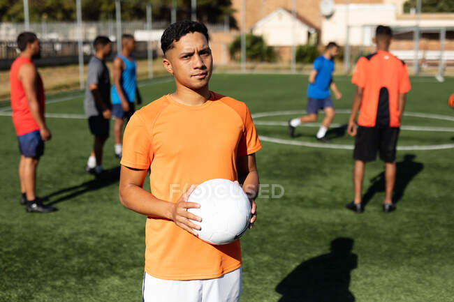 Portrait of focused mixed race male five a side football player wearing sports clothes training at a sports field in the sun, holding a ball his teammates warming up in the background. — Stock Photo