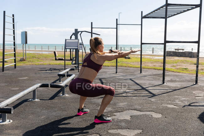 Vista lateral de una mujer atlética caucásica con pelo largo y oscuro haciendo ejercicio en un gimnasio al aire libre junto al mar durante el día, haciendo sentadillas. - foto de stock