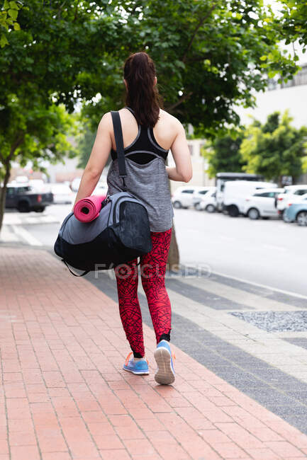 Передній вид підходящої кавказької жінки на шляху до тренування фітнесу в похмурий день, несучи спортивний мішок і кошик з йогою. — стокове фото