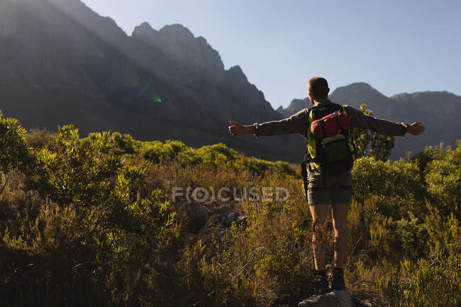 Rückansicht eines kaukasischen Mannes, der sich bei einem Ausflug in die Berge amüsiert, auf einem Feld unter den Bergen steht und die Aussicht genießt, die Arme weit ausgebreitet, an einem sonnigen Tag — Stockfoto