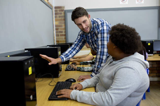 Side view of a male Caucasian teacher standing and talking with a teenage African American female high school student working on a computer and concentrating in a classroom — Stock Photo