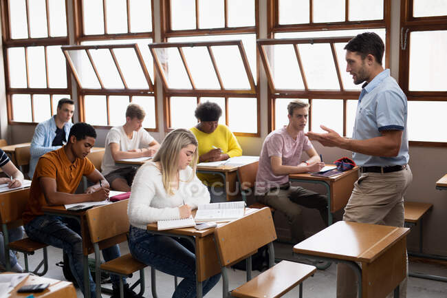 Vista lateral de un maestro de escuela secundaria masculino caucásico, de pie en el frente de una clase, hablando con un grupo multiétnico de adolescentes en un aula de la escuela sentado en escritorios en filas, escuchando - foto de stock
