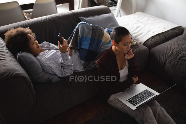 Vista ad alto angolo di una coppia mista di donne che si rilassano a casa in soggiorno, una sdraiata sul divano con una coperta sulle gambe usando uno smartphone, l'altra seduta accanto a lei sul pavimento usando un computer portatile — Foto stock