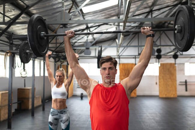 Front view of an athletic Caucasian man and woman wearing sports clothes cross training at a gym, standing and weight training with barbells, lifting the weights and holding them above their heads — Stock Photo