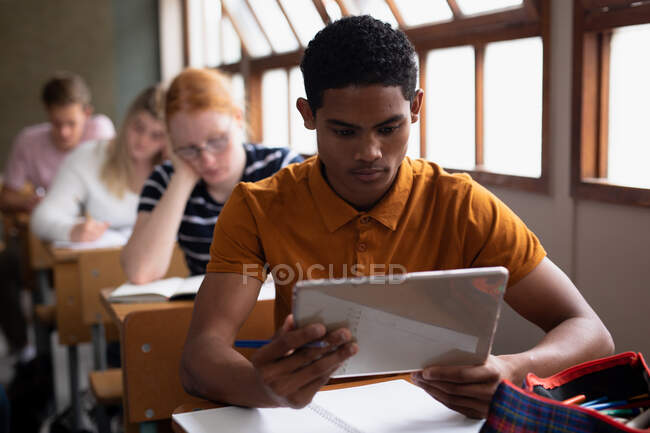 Front view close up of a teenage mixed-race boy in a school classroom sitting at desk, concentrating and using tablet computer, with teenage male and female classmates sitting at desks working in the background — Stock Photo