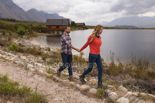 Vista lateral de una pareja caucásica pasándola bien en un viaje a las montañas, caminando por un sendero en una orilla del lago, tomados de la mano - foto de stock