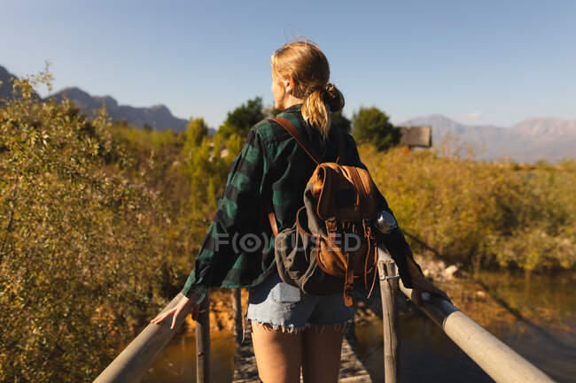 Вид сзади на кавказскую женщину, хорошо проводящую время в поездке в горы, стоящую на мосту, наслаждающуюся своим видом, в солнечный день — стоковое фото