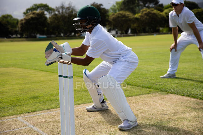 Side view of a teenage African American male cricket player wearing whites, helmet and gloves, standing on the pitch during a cricket match, waiting for a ball to catch, with other player standing in behind. — Stock Photo