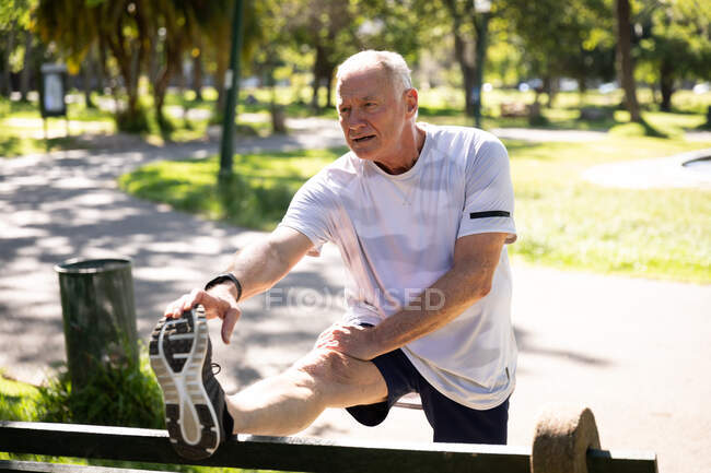 Front view of a mature senior Caucasian man working out in a park on a sunny day, stretching on a bench — Stock Photo