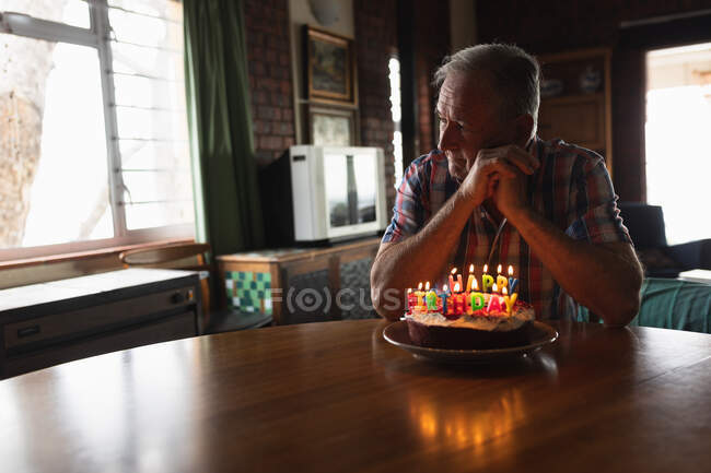Vista lateral de un hombre caucásico mayor en casa, sentado solo en la mesa del comedor con un pastel de cumpleaños con velas encendidas delante de él - foto de stock