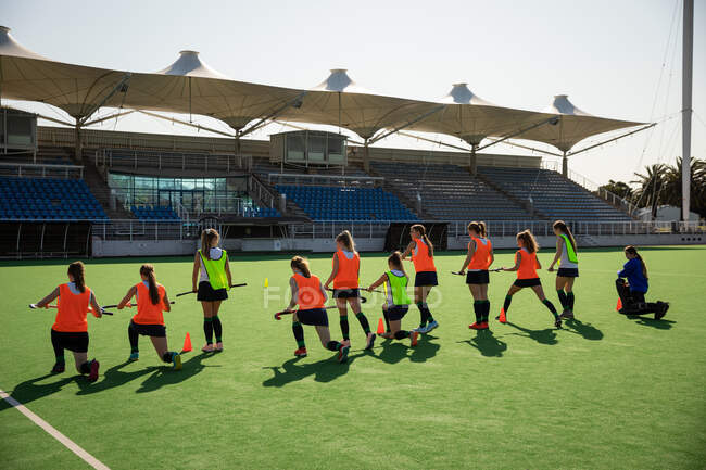 Side view of a group of female Caucasian field hockey players, training before a game, working out on a field hockey pitch, doing exercises, stretching their legs, holding hockey sticks on a sunny day — Stock Photo