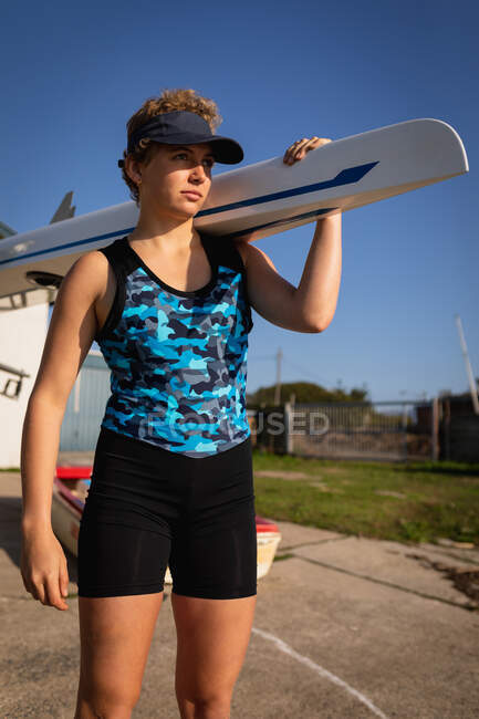 Nahaufnahme einer kaukasischen Ruderin mit Visier, die ein Boot auf der Schulter aus einem Bootshaus in der Sonne trägt, bevor sie auf dem Fluss trainiert — Stockfoto