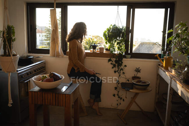 Кавказька жінка проводить час удома, ізолюючи і соціальну дистанцію в карантині під час епідемії коронавірусу (19), сидячи на кухні біля вікна і споживаючи каву.. — стокове фото