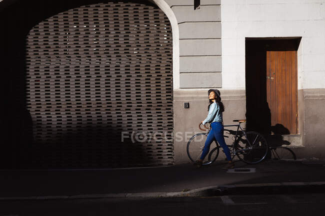 Seitenansicht einer fröhlichen Mischlingshündin mit langen dunklen Haaren, die tagsüber in den Straßen der Stadt unterwegs ist, einen Hut, Jeans und Jeansjacke trägt und mit ihrem Fahrrad mit Gebäude im Hintergrund spaziert. — Stockfoto