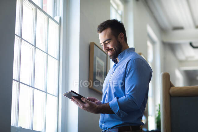Кавказский бизнесмен с короткими волосами, в голубой рубашке, работает в современном офисе, стоит у окна и пользуется планшетом — стоковое фото