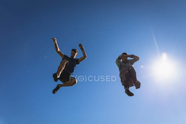 Front view of two Caucasian men practicing parkour by the building in a city on a cloudless sunny day, jumping up. — Stock Photo