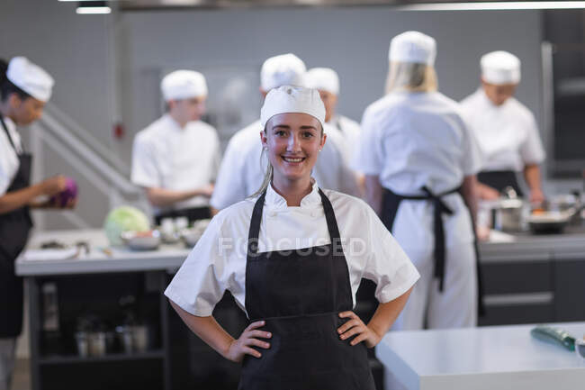 Porträt einer glücklichen kaukasischen Köchin, die in die Kamera blickt und die Hände lächelnd auf die Hüften legt, während andere Köche im Hintergrund kochen. Kochkurs in einer Restaurantküche Werkstatt kocht Essen. — Stockfoto