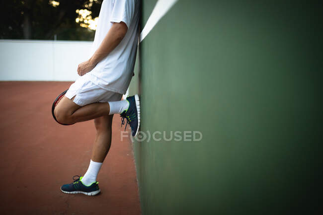 Вид чоловіка середньої секції в тенісному білому, який проводить час на корт, граючи в теніс в сонячний день, спираючись на стіну — стокове фото