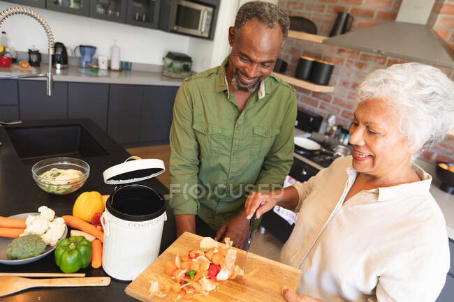 Високий кут зору щасливої пари афроамериканців похилого віку вдома, готувати їжу, зрізати овочі, покласти рослинні відходи в контейнер для компосту на своїй кухні, вдома разом ізолювати під час коронавірусної ковірусної ковідемії. — стокове фото