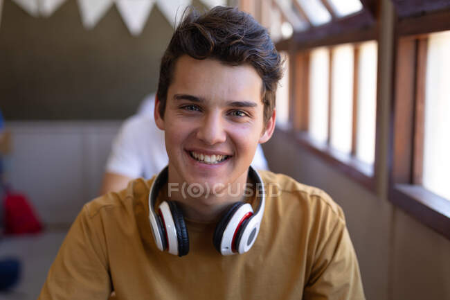 Retrato de cerca de un adolescente caucásico con cabello oscuro y ojos grises sentado en un escritorio en un aula de la escuela con auriculares alrededor de su cuello, sonriendo a la cámara - foto de stock