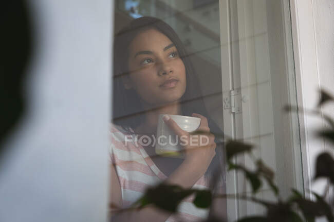 Mixed race woman spending time at home, standing and holding a mug. Self isolating and social distancing in quarantine lockdown during coronavirus covid 19 epidemic. — Stock Photo
